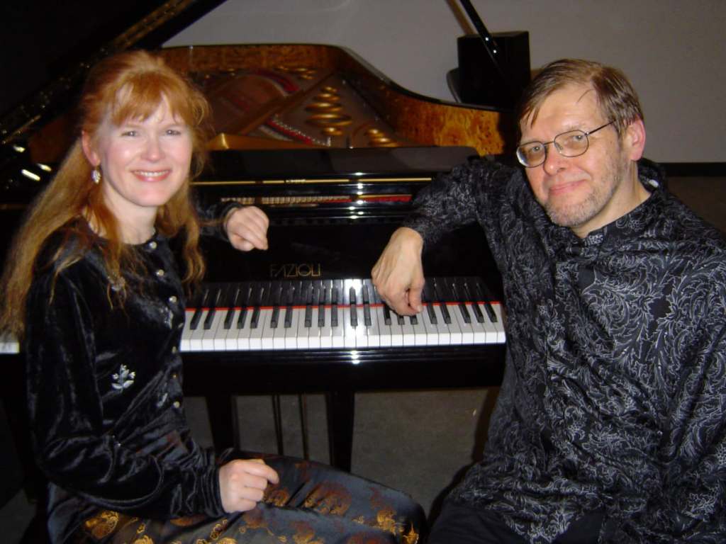 A red-haired woman and a man wearing glasses seated together at a piano.