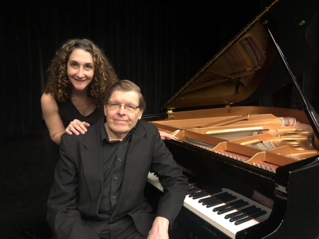 A man seated at a piano with a woman standing behind him with her hands on his shoulders.