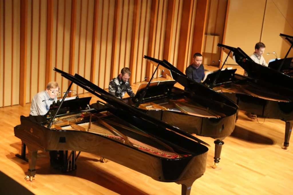 4 pianists playing 4 pianos lined up next to each other on a stage